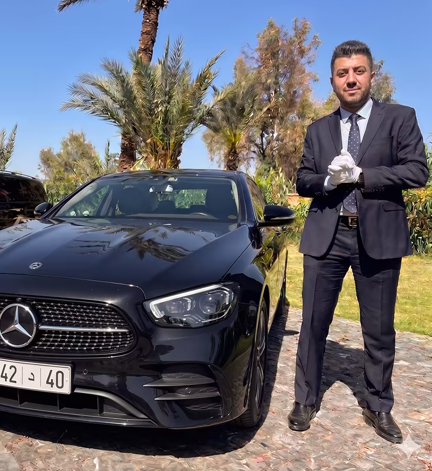 Chauffeur privé Northy en costume noir et gants blancs debout à côté d'une berline Mercedes Classe E noire haut de gamme sous le soleil de Tanger